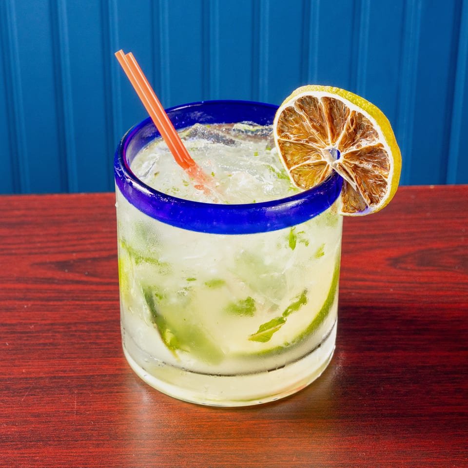 Iced lime and mint cocktail in a blue-rimmed glass with an orange straw and dried citrus garnish on a wooden table.