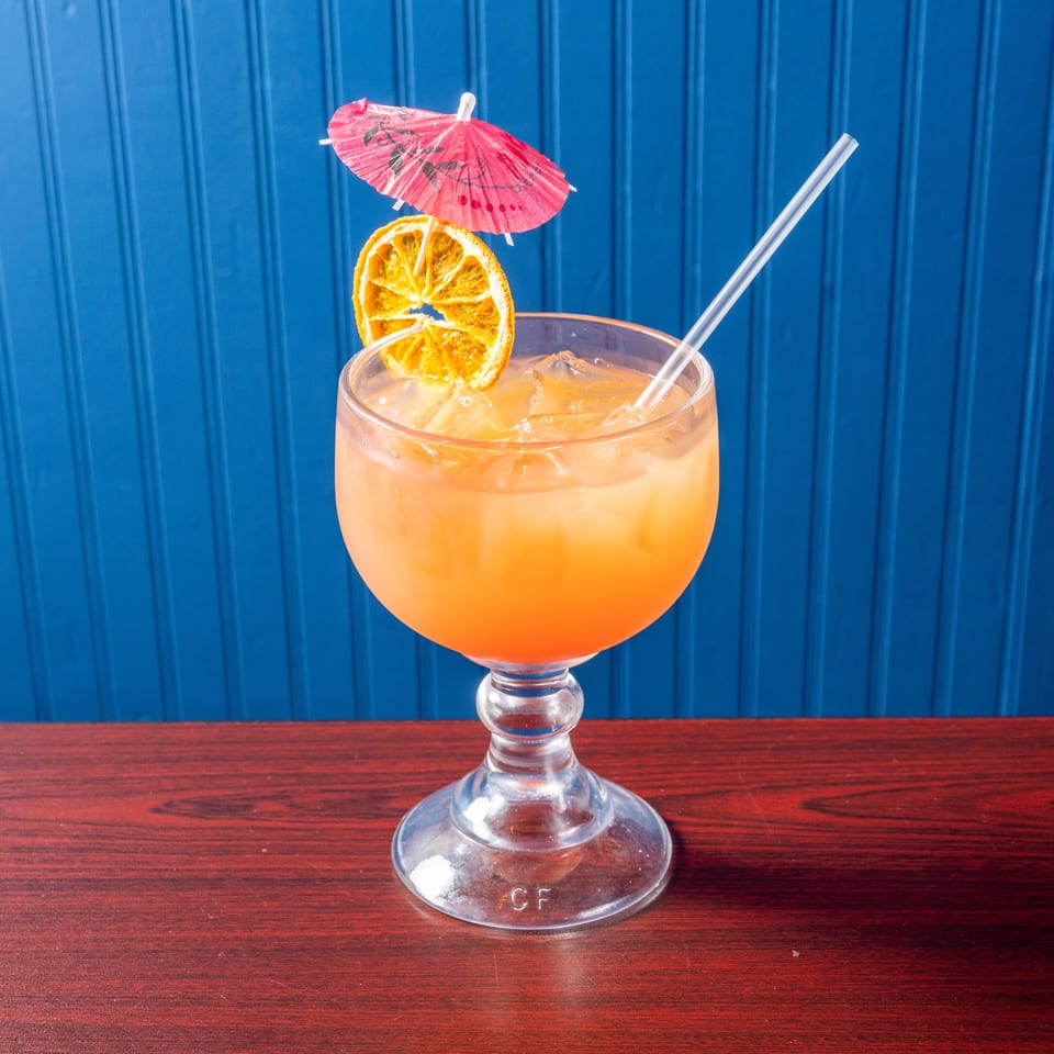 Orange cocktail with ice, straw, dried citrus slice, and pink paper umbrella in a stemmed glass on a wooden table.