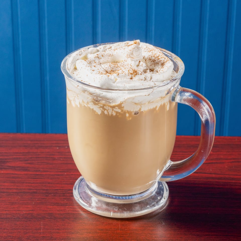 Creamy coffee topped with whipped cream and cinnamon served in a glass mug on a wooden table.