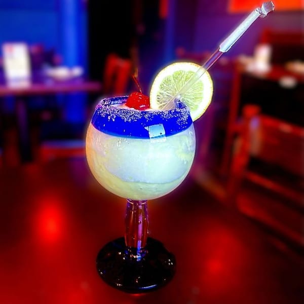 Frozen margarita with a blue rim, lemon slice, cherry, and straw in a restaurant setting.