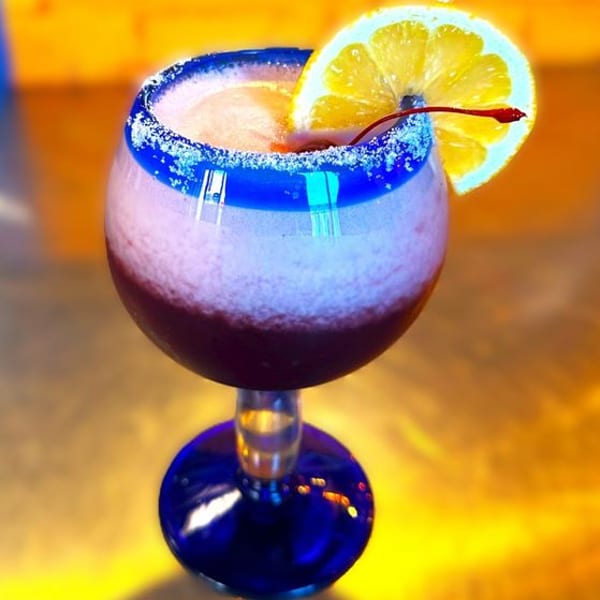 A vibrant, layered frozen drink in a blue-rimmed glass, garnished with a lemon slice and cherry.