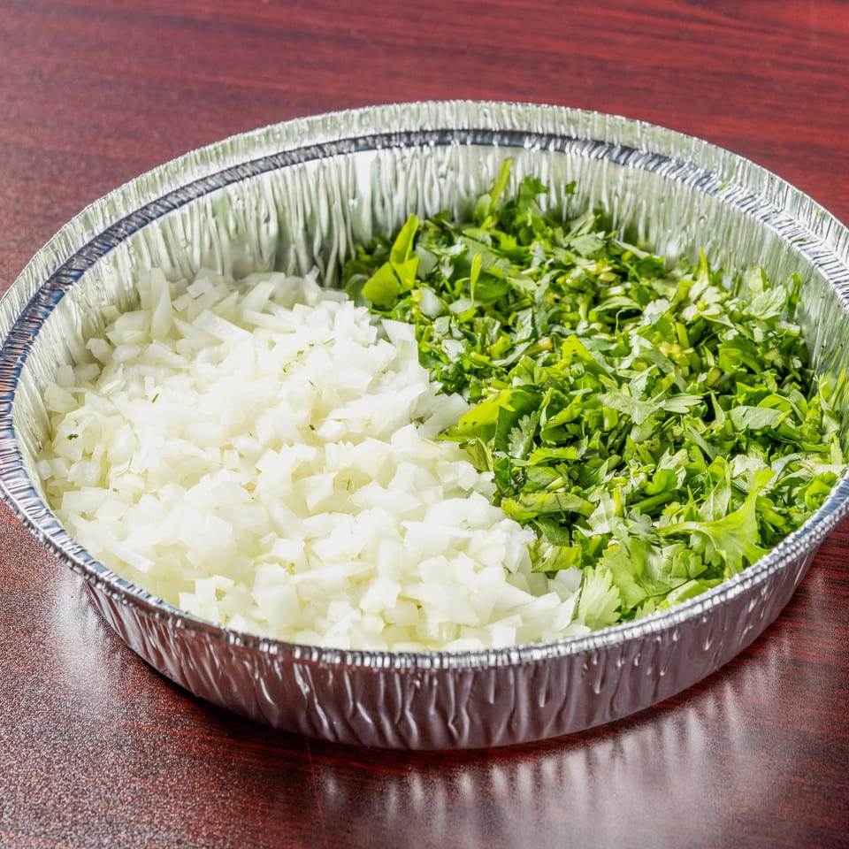 Aluminum tray filled with chopped white onions and fresh green herbs on a polished wooden table.
