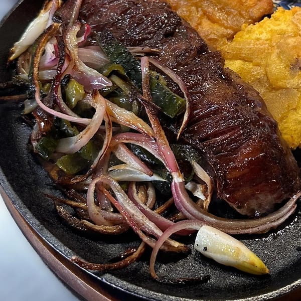 Sizzling steak with red onions, green peppers, and fried plantains on a black skillet.