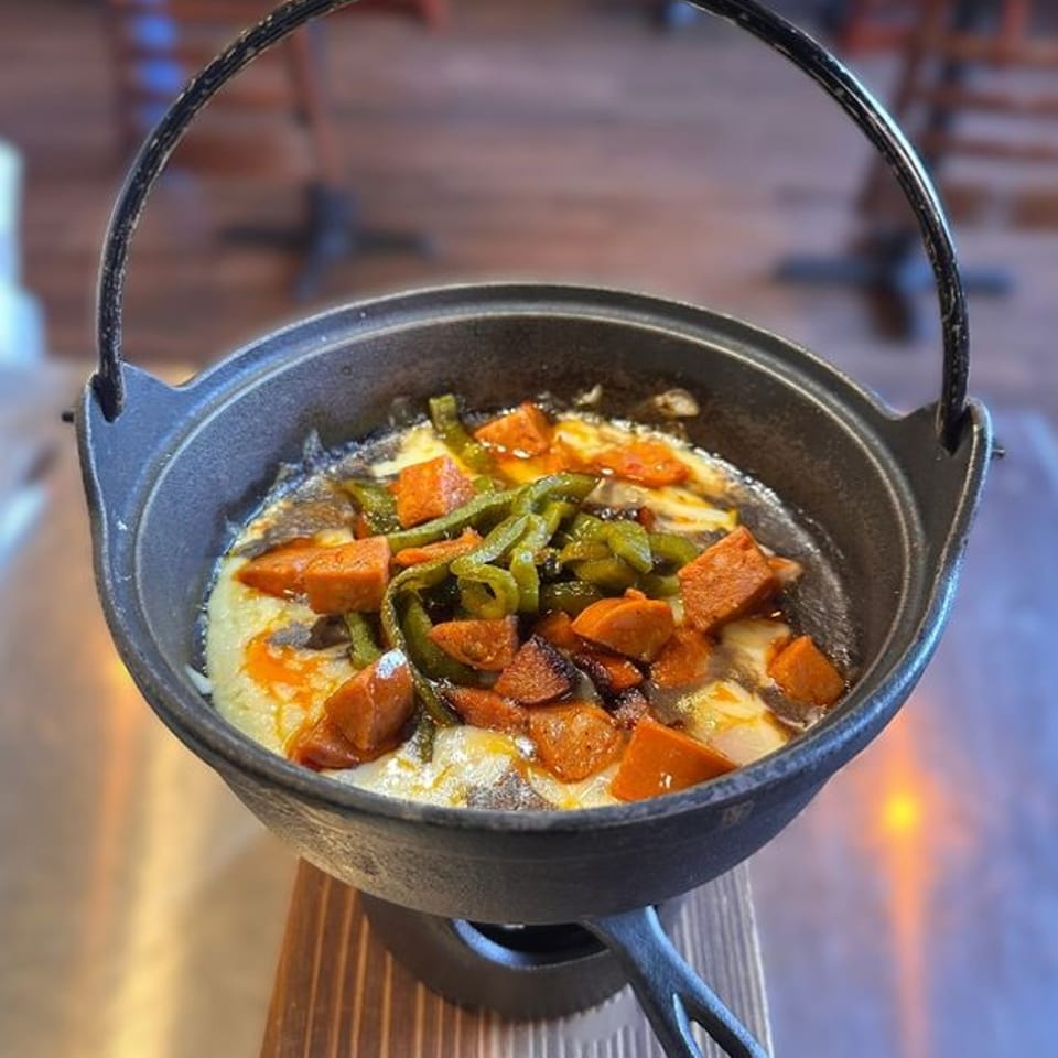 Melted cheese with chorizo and green peppers in a black cast iron pot on a wooden stand.