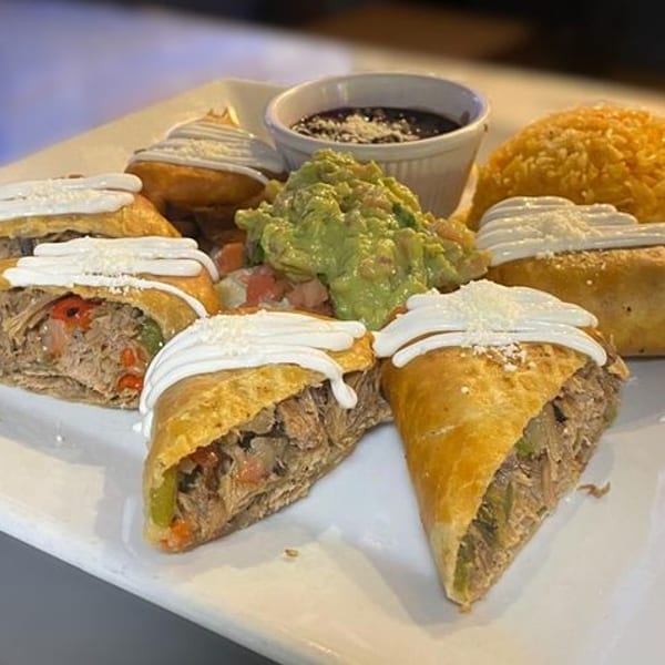 A white plate holds empanadas, rice, guacamole, and black beans, garnished with sour cream and cheese.