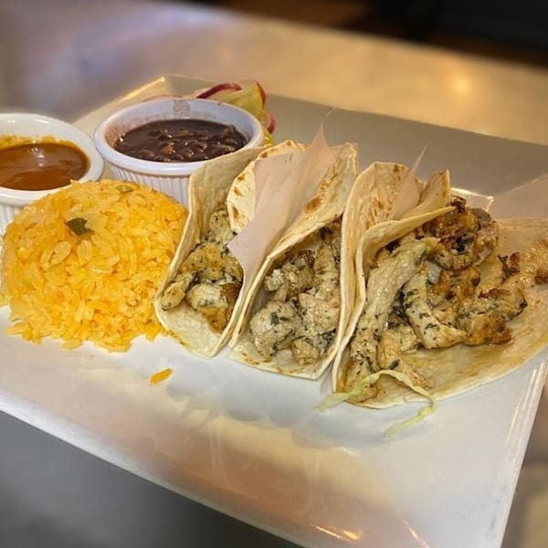 Three chicken tacos with rice, black beans, and salsa on a white rectangular plate.