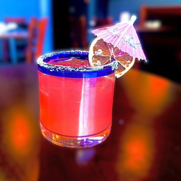 Bright red drink with a blue salt rim, orange slice, and pink umbrella on a dark table.