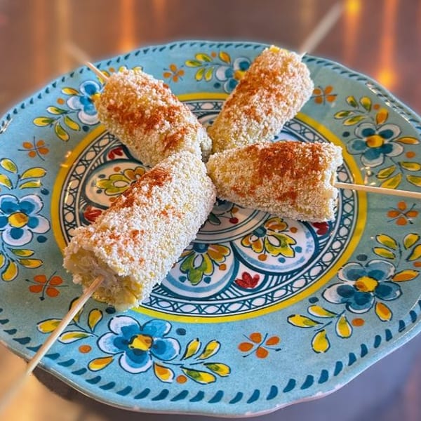 Four elote corn cobs on skewers, coated in white cheese and chili powder, on a colorful plate.